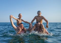 Zwei Väter und ihre Söhne genießen ein aufregendes Spiel im Meer, lachen, während sie auf den Schultern ihrer Väter balancieren; die Jungen halten sich triumphierend an den Händen und feiern ihren spielerischen Kampf