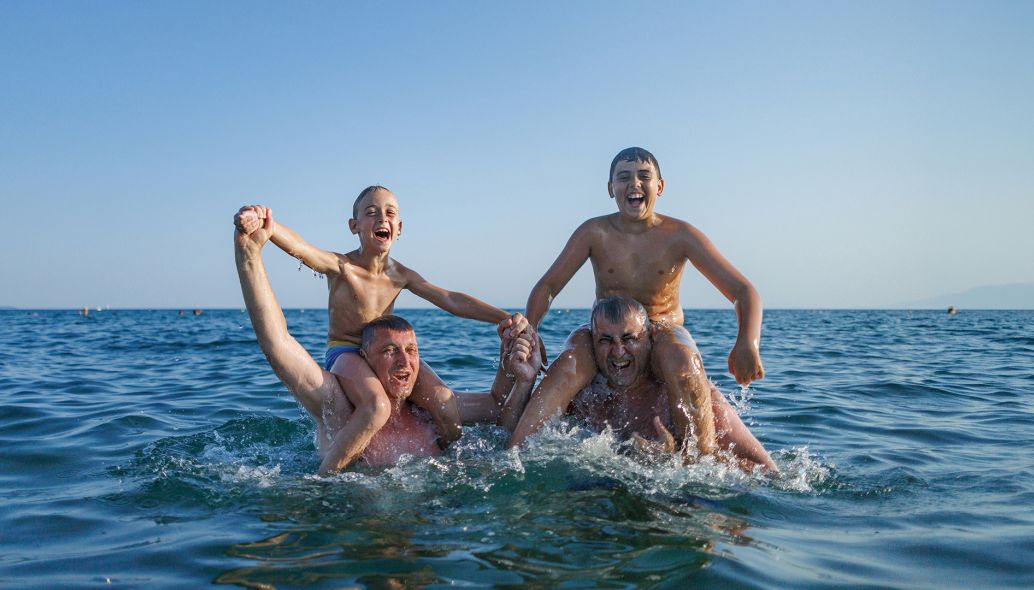 Zwei Väter und ihre Söhne genießen ein aufregendes Spiel im Meer, lachen, während sie auf den Schultern ihrer Väter balancieren; die Jungen halten sich triumphierend an den Händen und feiern ihren spielerischen Kampf