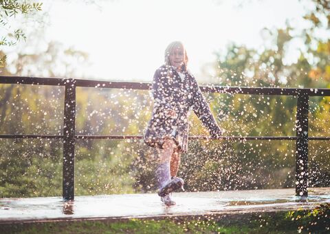 Ein Mädchen läuft lachend über eine nasse Holzbrücke und spritzt dabei Wasser auf. Die tief stehende Sonne scheint von hinten und taucht die Szene in warmes Licht.