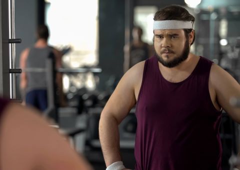 Ein junger Mann in Sportkleidung steht nachdenklich vor einem Spiegel im Fitnessstudio und schaut sich selbst an.