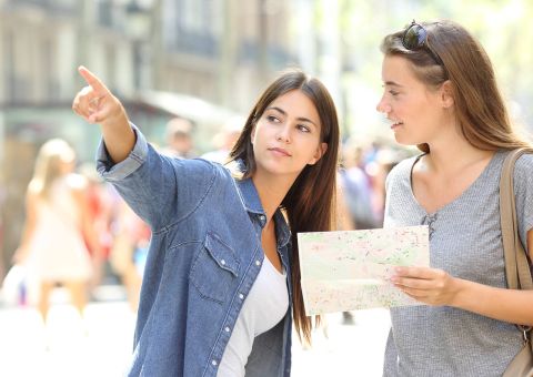 Zwei junge Frauen stehen beisammen – die eine deutet mit dem Finger in die Ferne, während die andere eine Landkarte in den Händen hält.