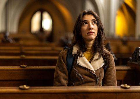 Eine junge Frau sitzt nachdenklich in einer leeren Kirche und blickt mit nachdenklich, fragender Miene nach oben.