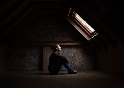 Frau sitzt nachdenklich vor einem Dachfenster auf dem Boden eines Dachbodens.