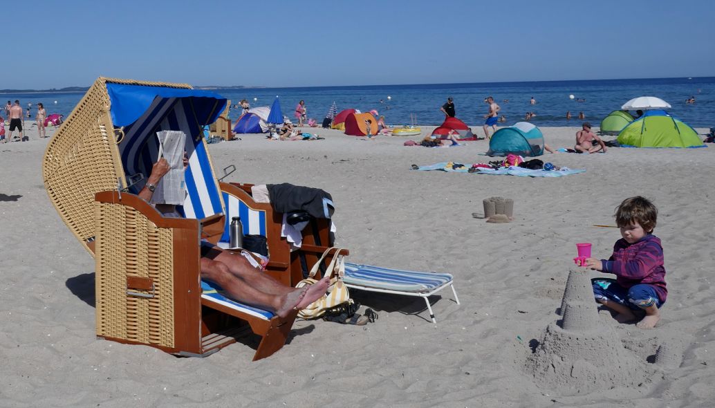 Mann im Strandkorb auf Rügen an der Ostsee mit einem Kind das eine Sandburg baut. KI generiert.