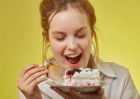 Junge Frau freut sich über ein Stück Sahnetorte mit Beeren auf gelbem Hintergrund.