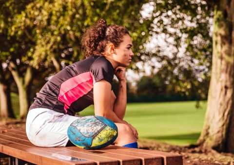 Eine junge Sportlerin sind nachdenklich auf der Bank am Rande des Sportplatzes. Neben ihr liegt ein Rugbyball.